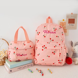 May include: A pink backpack and matching lunch bag, both adorned with a cherry pattern. The backpack features the name "Vivian" embroidered in pink. The lunch bag also has the name "Vivian" embroidered on it. The bags are set on a white surface.