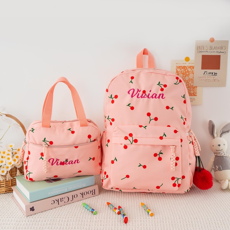 May include: A pink backpack and matching lunch bag, both adorned with a cherry pattern. The backpack features the name "Vivian" embroidered in pink. The lunch bag also has the name "Vivian" embroidered on it. The bags are set on a white surface.
