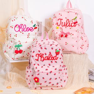 May include: Three corduroy backpacks with cherry and strawberry patterns. One is white with "Chloe" embroidered, another is pink with "Julia", and the third is pink with "Thalida". Each has a front pocket and a top handle.