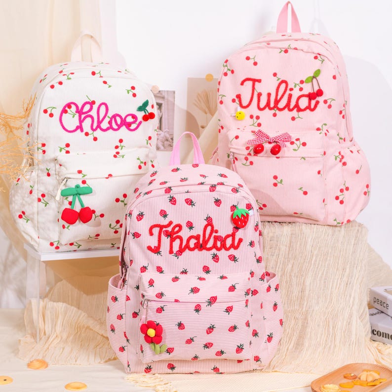 May include: Three corduroy backpacks with cherry and strawberry patterns. One is white with "Chloe" embroidered, another is pink with "Julia", and the third is pink with "Thalida". Each has a front pocket and a top handle.