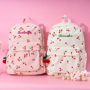 May include: Two white canvas cherry print backpacks with red cherries. The backpack on the left is pink and says "Isabella" in pink. The backpack on the right is white and says "Natasha" in green.