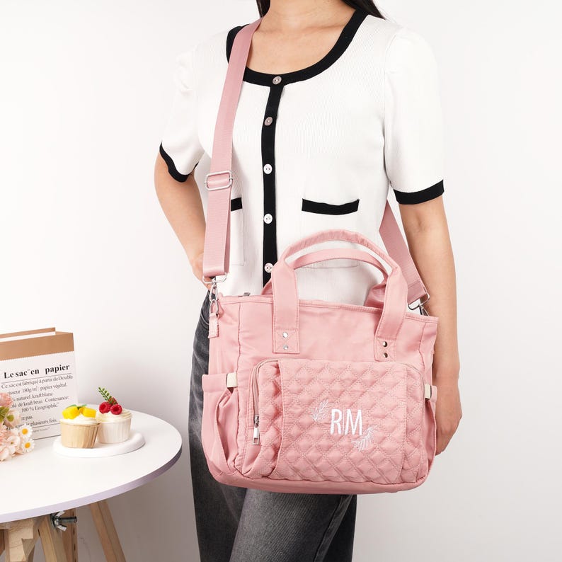 May include: A pink quilted tote bag with a shoulder strap and top handles. The bag has a front pocket with the embroidered initials "RM". The bag is made of a durable, water-resistant material and has multiple compartments for storage.