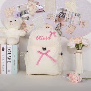 May include: Cream-colored backpack with the name "Olivia" embroidered in pink. The backpack features pink bows and a small bear-shaped clasp. A white teddy bear and books are visible in the background.