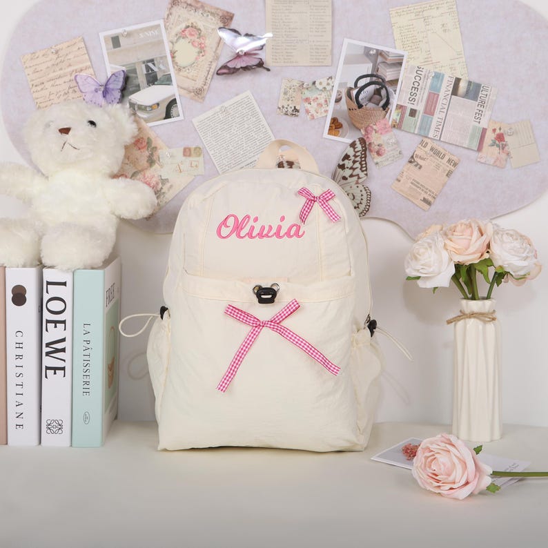 May include: Cream-colored backpack with the name "Olivia" embroidered in pink. The backpack features pink bows and a small bear-shaped clasp. A white teddy bear and books are visible in the background.