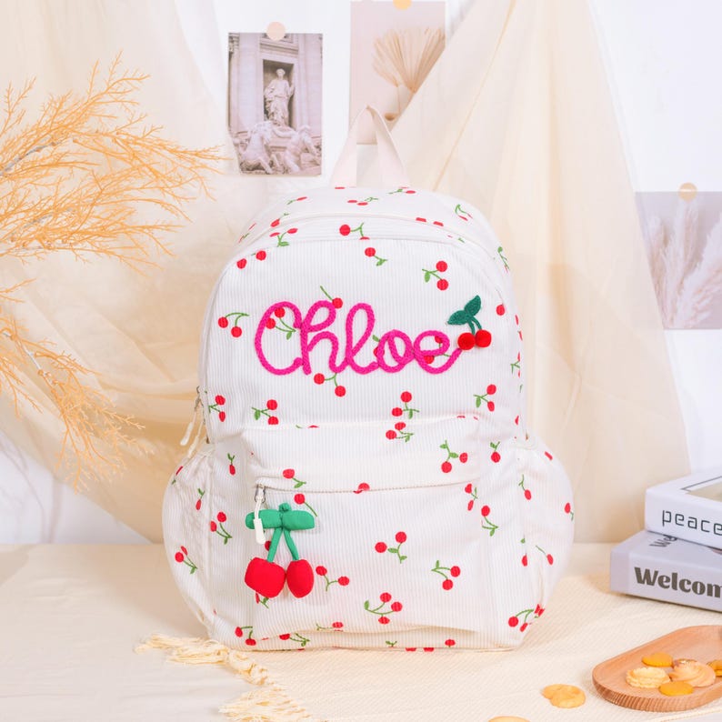 May include: White corduroy backpack with a cherry pattern and the name "Chloe" embroidered in pink. Features a front pocket with a cherry charm and a top handle. The backpack is decorated with red cherries and green leaves.