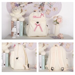 May include: Cream-colored backpack with the name "Olivia" embroidered in pink. The backpack features a front pocket with a pink bow and adjustable straps. A teddy bear, books, and a small lamp are visible in the background.
