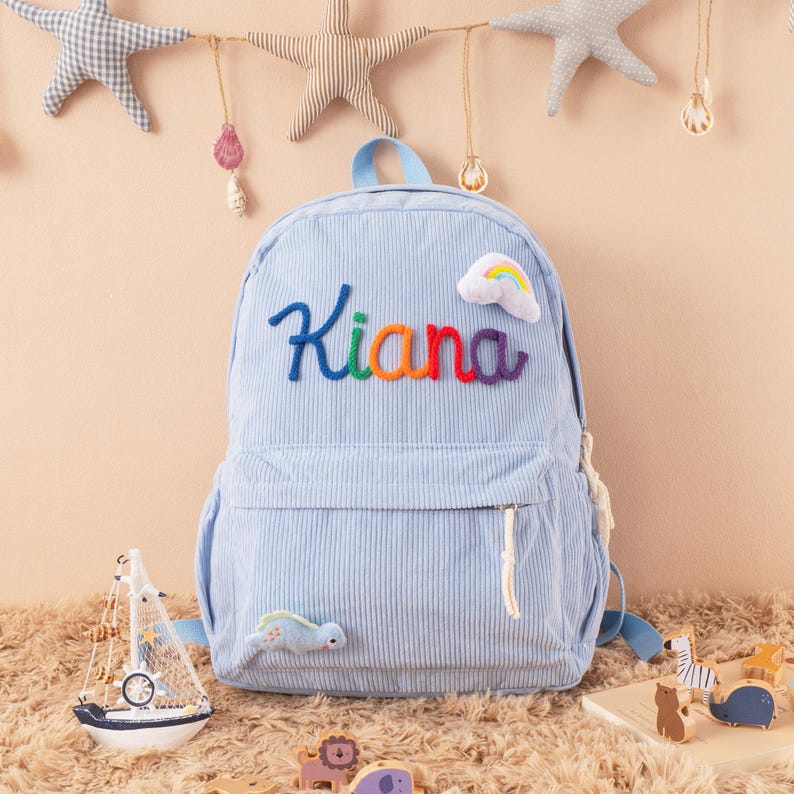 May include: Light blue corduroy backpack with the name "Kiana" embroidered in colorful thread. A small rainbow and a dinosaur patch are also on the backpack. The backpack has a top handle and adjustable straps.