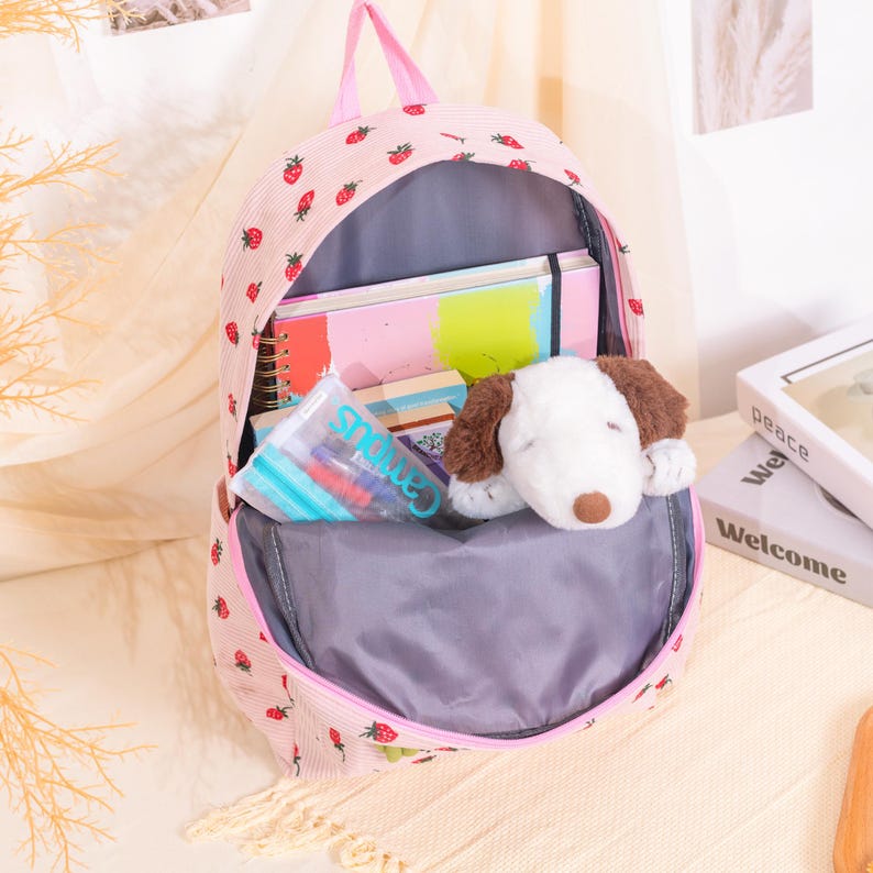 May include: Pink backpack with a strawberry pattern, open to reveal school supplies and a plush dog toy. The backpack has a pink handle and trim. Inside are notebooks, pens, and a stuffed animal.