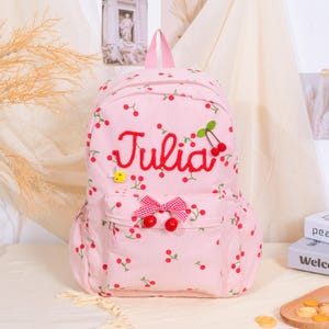 May include: Pink corduroy backpack with a cherry pattern and the name "Julia" embroidered in red. Features a front pocket with a bow and cherry accents. Includes a top handle and adjustable straps.