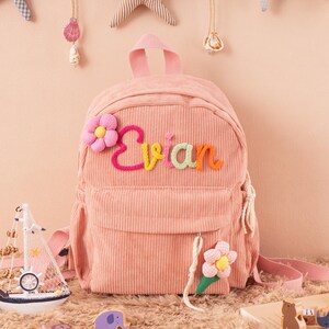 May include: A pink corduroy backpack with the name "Evian" embroidered in colorful letters. The backpack features a flower applique and a small front pocket. A small sailboat and other decorative items are in the background.