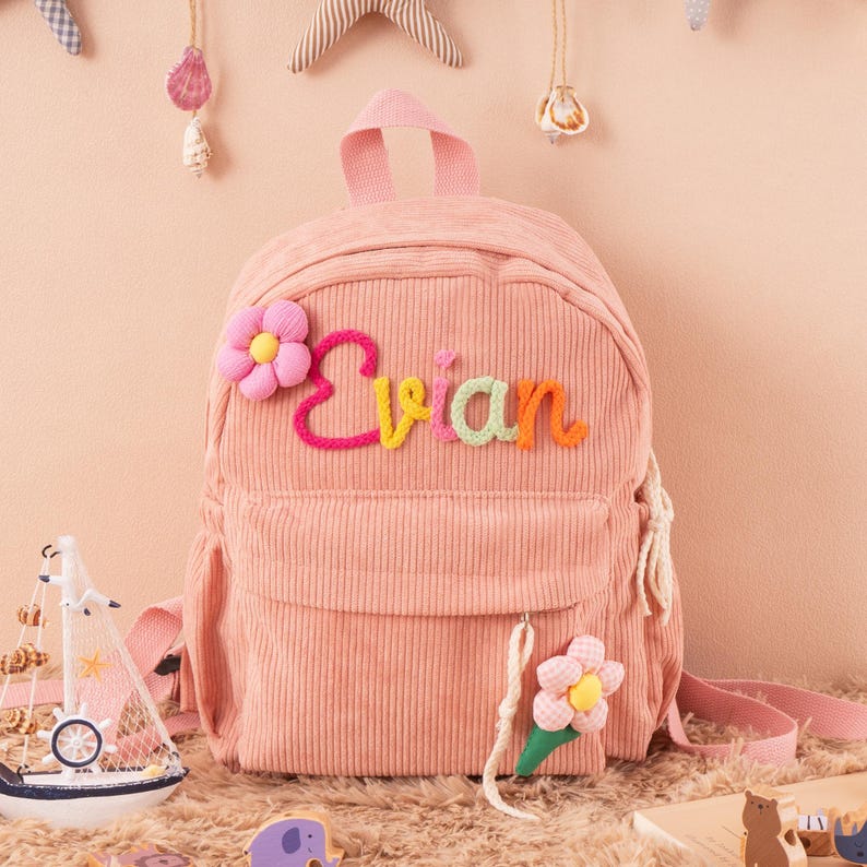 May include: A pink corduroy backpack with the name "Evian" embroidered in colorful letters. The backpack features a flower applique and a small front pocket. A small sailboat and other decorative items are in the background.