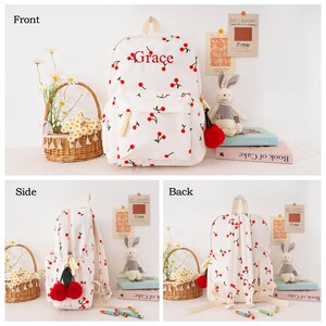 May include: White backpack with a cherry pattern and the name "Grace" in red script. The backpack has a front pocket, a top handle, and adjustable straps. The zipper pull has a red pom-pom.