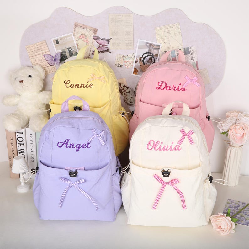 May include: Four pastel-colored backpacks are displayed, each with a name embroidered on the front. The backpacks are yellow, pink, lavender, and white, with matching bows and accents. The names embroidered are "Connie", "Doris", "Angel", and "Olivia".