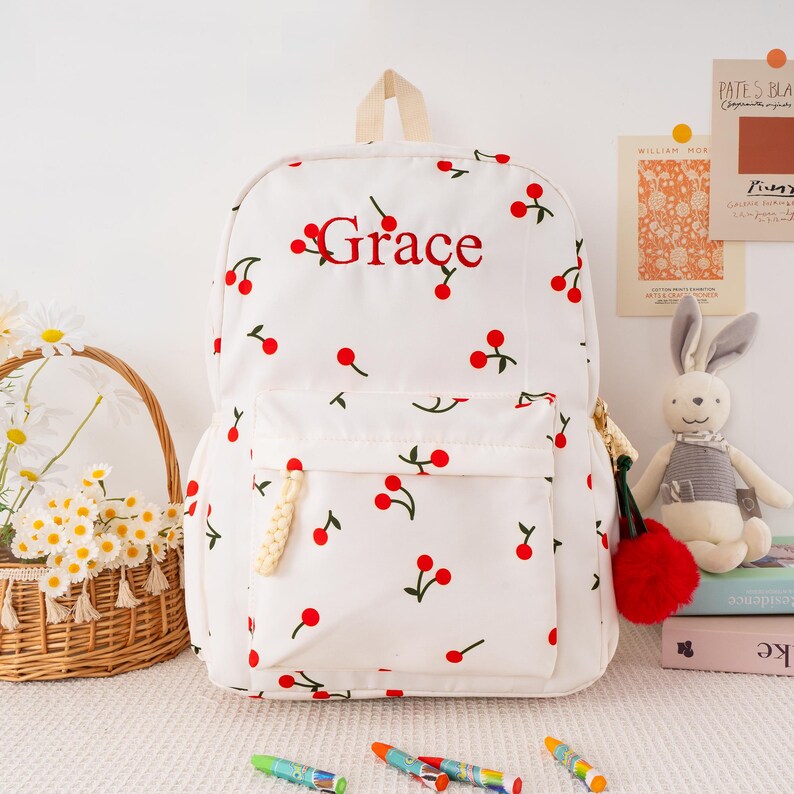 May include: White backpack with a cherry pattern and the name "Grace" embroidered in red. The backpack has a front pocket, a top handle, and a red pom-pom charm. A basket of daisies and a stuffed bunny are in the background.