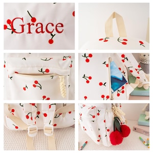 May include: White backpack with a cherry pattern and the name "Grace" embroidered in red. The backpack features a front pocket, adjustable straps, and a top handle. A red pom-pom cherry charm hangs from the zipper.