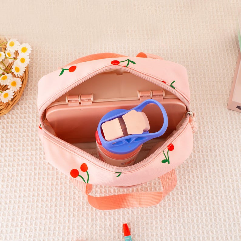 May include: A pink lunch bag with a cherry pattern, open to reveal a pink lunchbox and a blue and pink water bottle. The bag has a matching pink handle and is set on a white, textured surface.