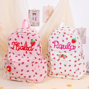 May include: Two corduroy backpacks, one pink and one white, are covered in small strawberry patterns. The pink backpack has the name "Thalida" embroidered in red, and the white backpack has the name "Paula" embroidered in pink.