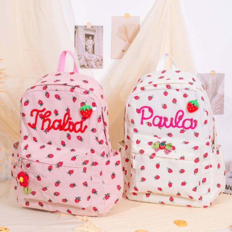 May include: Two corduroy backpacks, one pink and one white, are covered in small strawberry patterns. The pink backpack has the name "Thalida" embroidered in red, and the white backpack has the name "Paula" embroidered in pink.