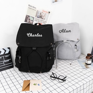 May include: Two black and gray canvas backpacks with adjustable straps. The black backpack has the name "Charles" embroidered on the front. The gray backpack has the name "Alan" embroidered on the front.