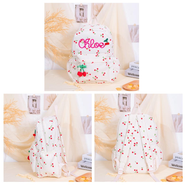 May include: White backpack with a cherry pattern and the name "Chloe" embroidered in pink. The backpack features a front pocket and a 3D cherry embellishment. The bag has adjustable straps and a top handle.
