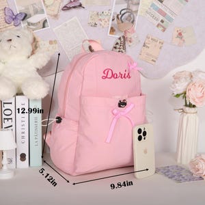 May include: A pink backpack with the name "Doris" embroidered on it. The backpack has a front pocket and bow accents. Dimensions are 12.99 inches high, 9.84 inches wide, and 5.12 inches deep. A phone rests beside it.