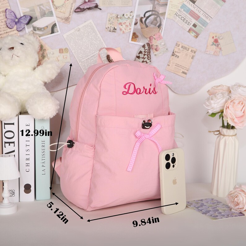 May include: A pink backpack with the name "Doris" embroidered on it. The backpack has a front pocket and bow accents. Dimensions are 12.99 inches high, 9.84 inches wide, and 5.12 inches deep. A phone rests beside it.