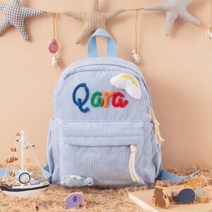 May include: Light blue corduroy backpack with the name "Qara" in colorful letters and a rainbow cloud applique. The backpack has a front pocket, a top handle, and adjustable straps. Decorative nautical-themed toys are in the foreground.