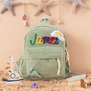 May include: A light green corduroy backpack with the name "James" embroidered in colorful letters. The backpack features a small front pocket, a rainbow and cloud patch, and a small dinosaur applique. Decorative stars and a sailboat are in the background.