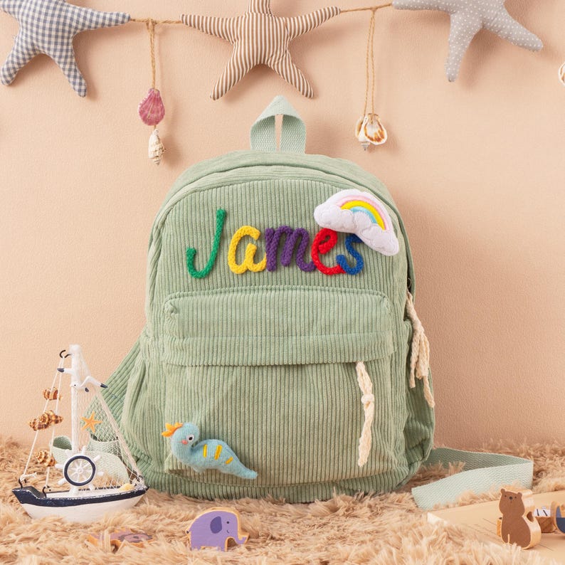 May include: A light green corduroy backpack with the name "James" embroidered in colorful letters. The backpack features a small front pocket, a rainbow and cloud patch, and a small dinosaur applique. Decorative stars and a sailboat are in the background.
