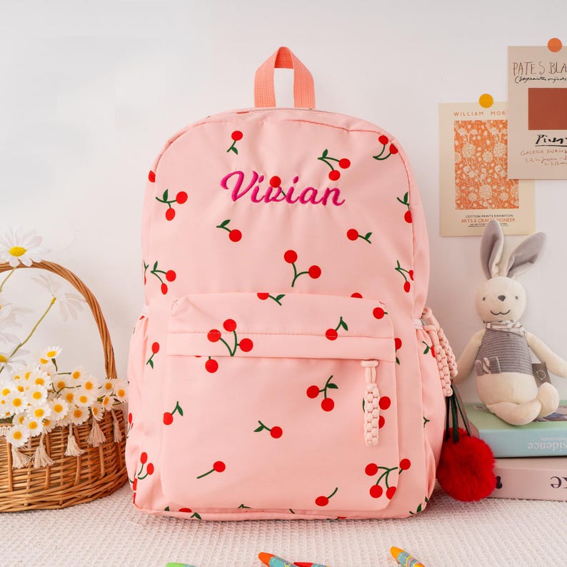 May include: Pink backpack with a cherry pattern and the name "Visian" embroidered in pink. The backpack has a front pocket, a top handle, and a red pom-pom zipper pull. A cute and stylish accessory for everyday use.