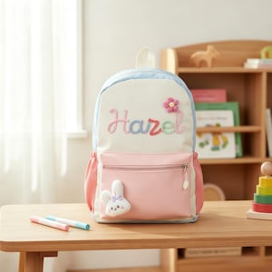 May include: A cream-coloured backpack with light blue trim and a pink front pocket. The name Hazel is embroidered in multicoloured script above a pink flower appliqu&eacute;. A small white plush rabbit with a purple bow is attached to the front pocket.