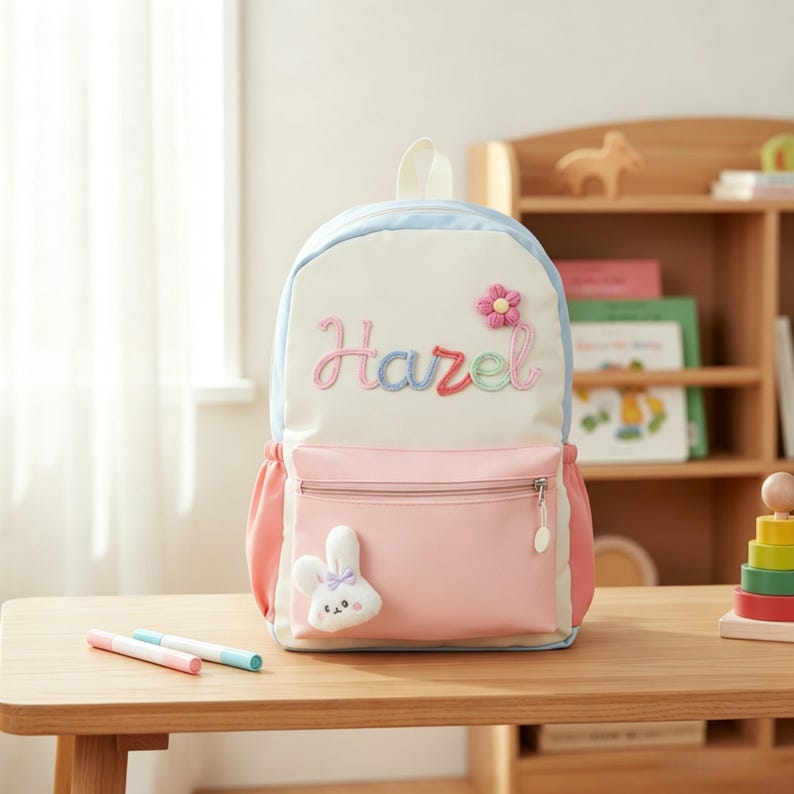 May include: A cream-coloured backpack with light blue trim and a pink front pocket. The name Hazel is embroidered in multicoloured script above a pink flower appliqu&eacute;. A small white plush rabbit with a purple bow is attached to the front pocket.