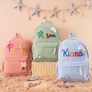 May include: Three corduroy backpacks in pink, green, and blue, each with a name embroidered in colorful thread. The pink backpack says "Paula", the green says "Mason", and the blue says "Kiana". Decorative elements include flowers, clouds, and a bird.