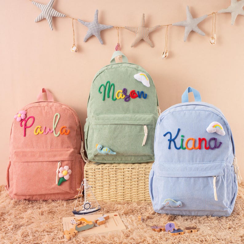 May include: Three corduroy backpacks in pink, green, and blue, each with a name embroidered in colorful thread. The pink backpack says "Paula", the green says "Mason", and the blue says "Kiana". Decorative elements include flowers, clouds, and a bird.