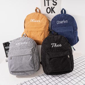 May include: Four corduroy backpacks in different colors with personalized names embroidered on the front: Katelyn, Oliver, Theo, and Charles.