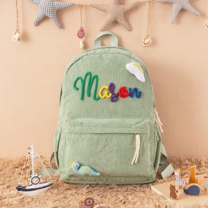 May include: A light green corduroy backpack with the name "Mason" embroidered in colorful thread. The backpack has a front pocket, a top handle, and a small rainbow and dinosaur applique. Decorative stars and a toy sailboat are in the background.