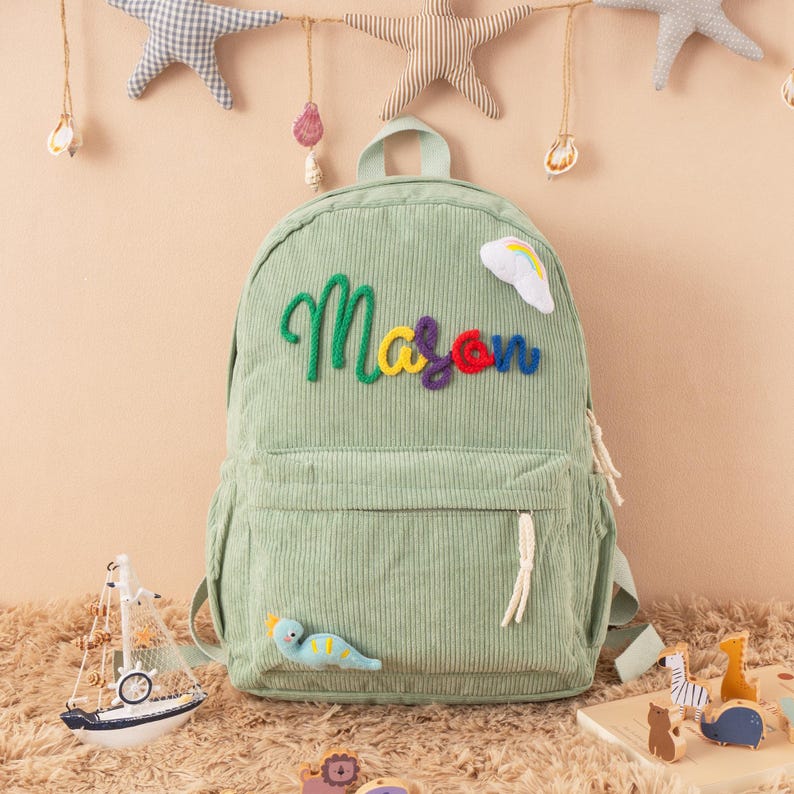 May include: A light green corduroy backpack with the name "Mason" embroidered in colorful thread. The backpack has a front pocket, a top handle, and a small rainbow and dinosaur applique. Decorative stars and a toy sailboat are in the background.