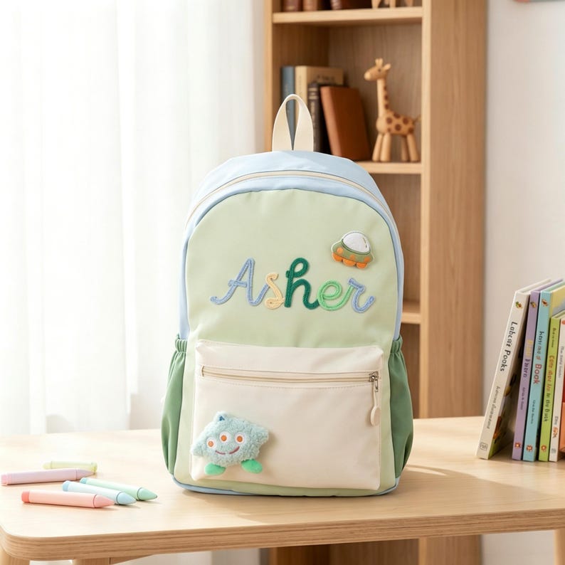 May include: A light green and cream backpack featuring the embroidered name Asher in blue and green script. The bag includes a small plush alien character attached to the front pocket, a small UFO patch, a top handle, and side pockets.