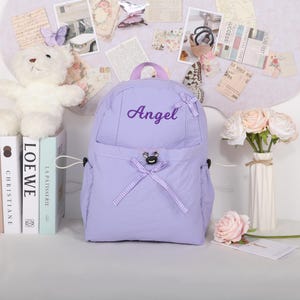 May include: A lavender backpack with the name "Angel" embroidered on the front. The backpack has a top handle, two side pockets, and a front pocket with a bow. A white teddy bear and books are on the left side of the backpack.
