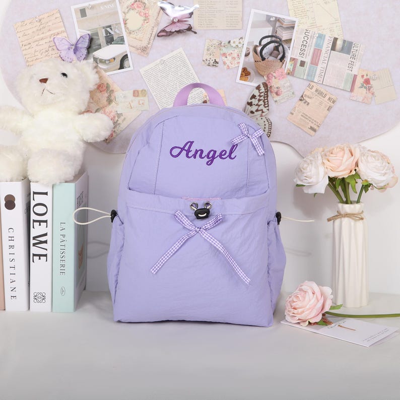 May include: A lavender backpack with the name "Angel" embroidered on the front. The backpack has a top handle, two side pockets, and a front pocket with a bow. A white teddy bear and books are on the left side of the backpack.