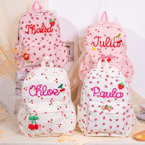 May include: Four backpacks with strawberry and cherry patterns. Two are pink, two are white. Each backpack has a name embroidered on it: Thalod, Julia, Chloe, and Paula. The backpacks have front pockets and side pockets.