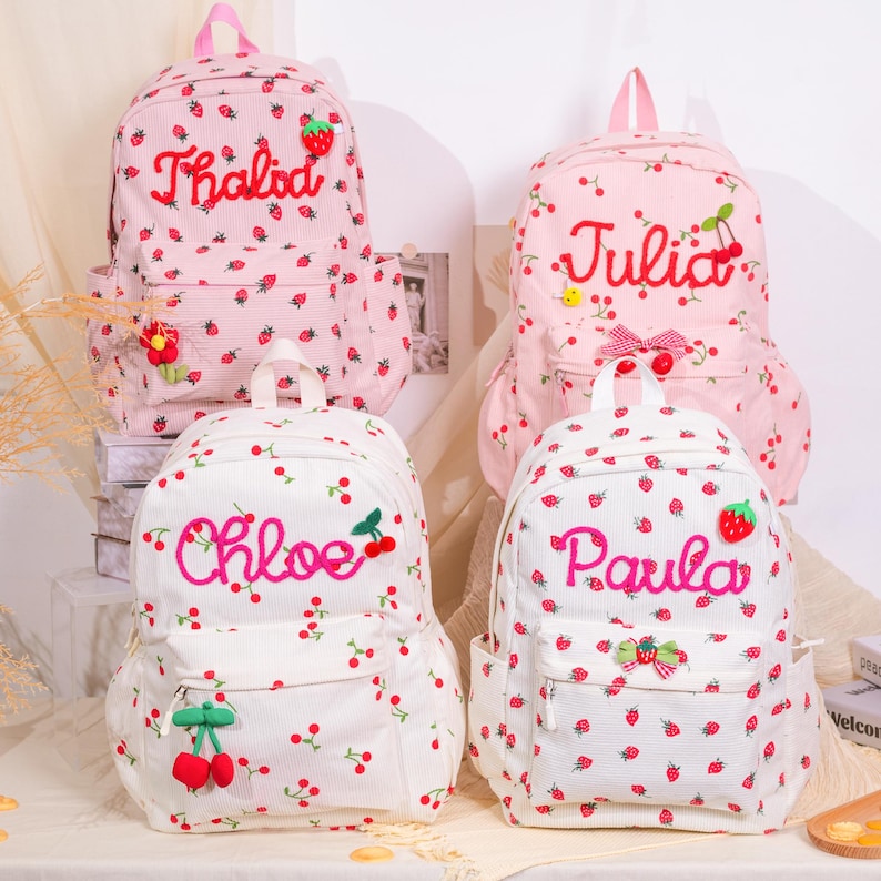 May include: Four backpacks with strawberry and cherry patterns. Two are pink, two are white. Each backpack has a name embroidered on it: Thalod, Julia, Chloe, and Paula. The backpacks have front pockets and side pockets.