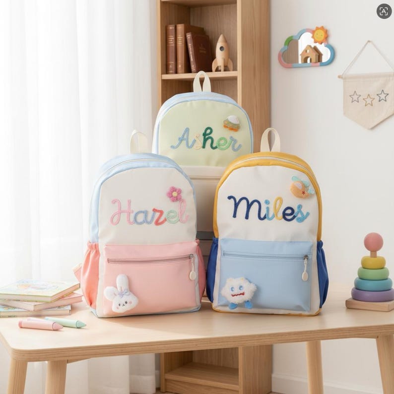 May include: Three backpacks with personalised names embroidered on the front. The left bag is white and pink with a bunny patch, the centre bag is light green with a rocket patch, and the right bag is white and blue with a cloud patch.