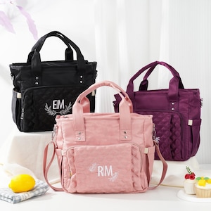 May include: Three diaper bags in black, pink, and purple. Each bag has a top handle, a shoulder strap, and multiple pockets. The black bag has "EM" embroidered on it, the pink bag has "RM" embroidered on it. All bags have a quilted pattern.