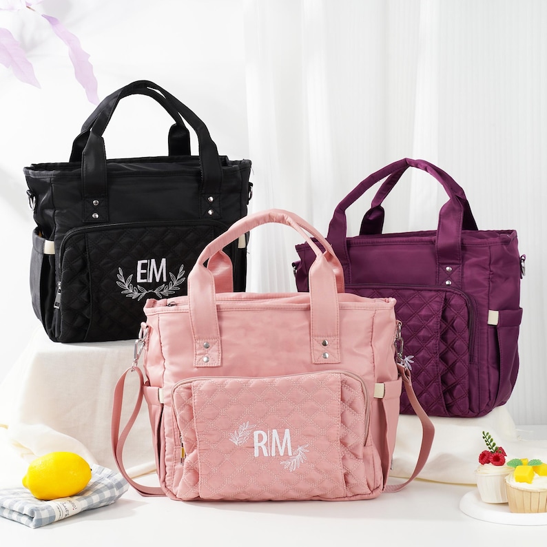 May include: Three diaper bags in black, pink, and purple. Each bag has a top handle, a shoulder strap, and multiple pockets. The black bag has "EM" embroidered on it, the pink bag has "RM" embroidered on it. All bags have a quilted pattern.