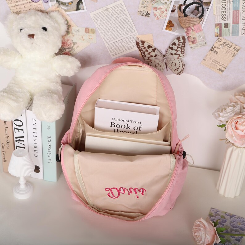 May include: A pink backpack with the name "Doris" embroidered in cursive. The backpack is open, revealing books and interior pockets. A white teddy bear and books are on the left. The text "National Trust Book of Bread" is visible.