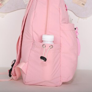 May include: A light pink backpack with a water bottle in the side pocket. The backpack has a zipper closure and adjustable straps. A white water bottle is in the side pocket. The backpack has a small pocket on the front with a pink bow.