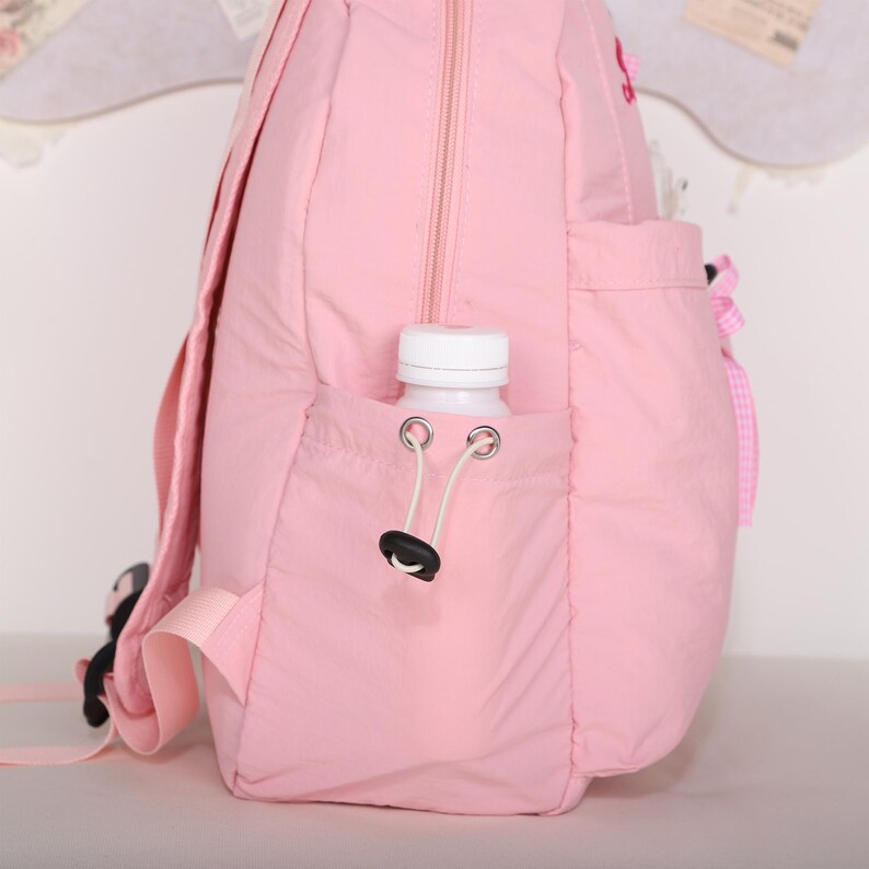 May include: A light pink backpack with a water bottle in the side pocket. The backpack has a zipper closure and adjustable straps. A white water bottle is in the side pocket. The backpack has a small pocket on the front with a pink bow.