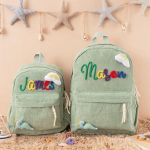 May include: Two green corduroy backpacks, one smaller than the other. The smaller backpack has the name "James" embroidered in colorful thread, and the larger one says "Mason". Each has a rainbow and a dinosaur applique.