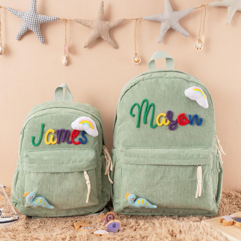 May include: Two green corduroy backpacks, one smaller than the other. The smaller backpack has the name "James" embroidered in colorful thread, and the larger one says "Mason". Each has a rainbow and a dinosaur applique.